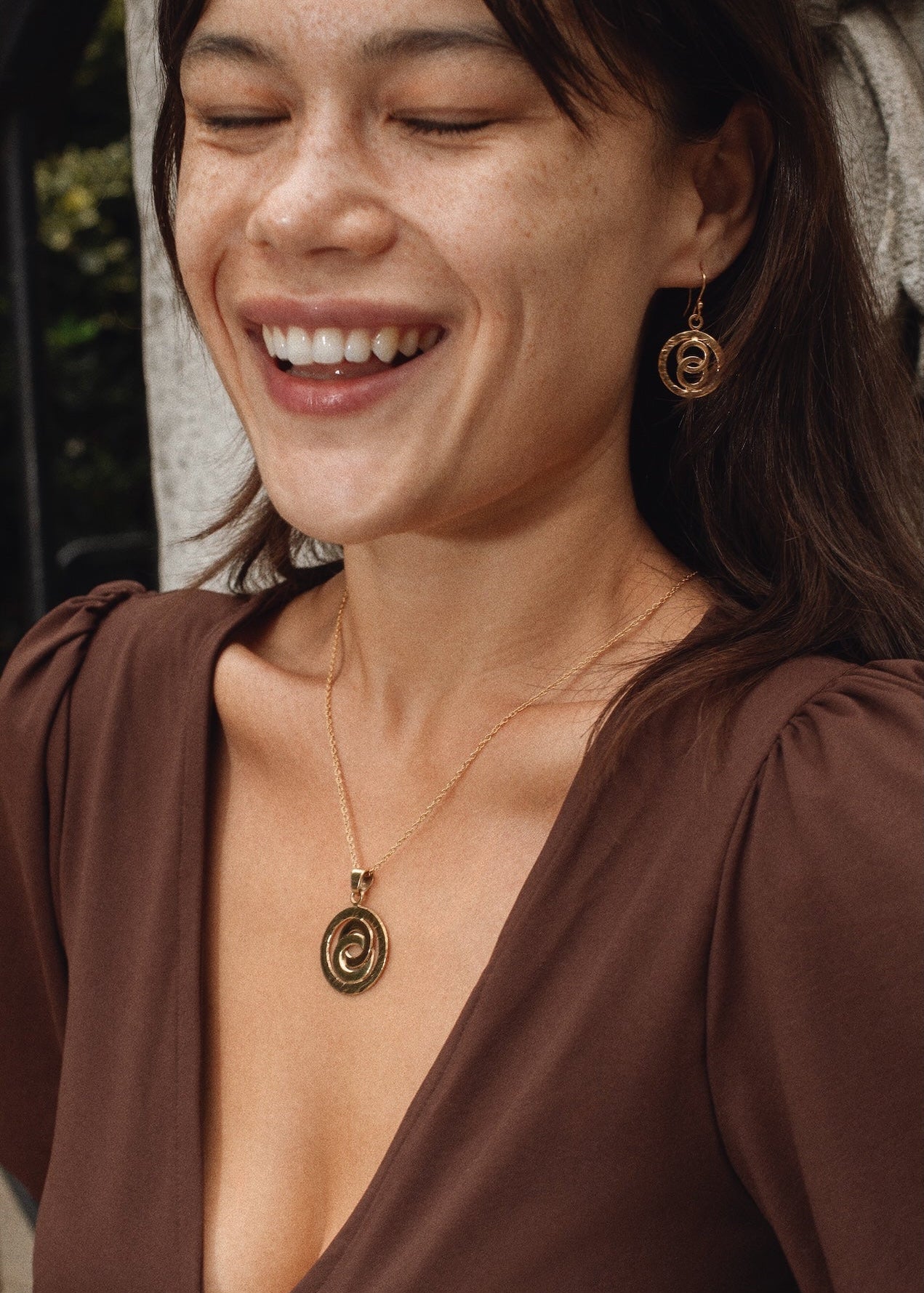 Woman wearing a gold necklace and earrings, smiling outdoors.