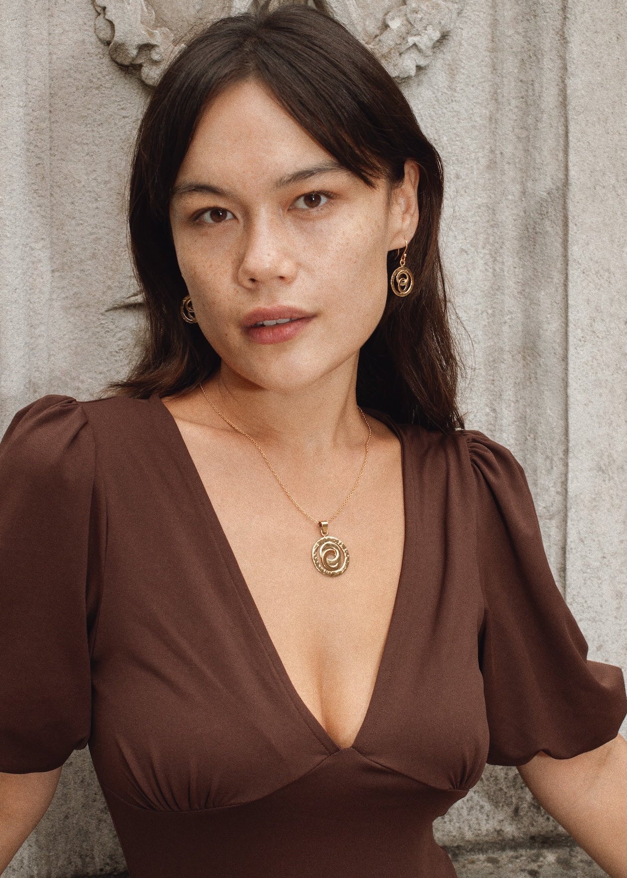 Woman wearing a brown dress with a deep neckline, standing against a textured wall.