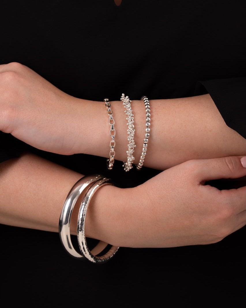 A close-up of a person's crossed arms wearing several silver-toned bracelets, including a delicate beaded chain bracelet, a textured link bracelet, and two polished bangle bracelets, against a black top and black background.