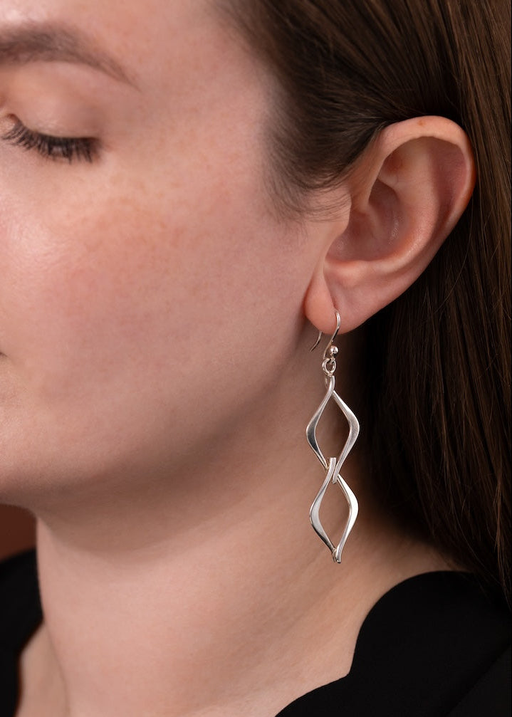 A close-up of a woman wearing a silver dangle earring with a twisted, spiral design. The earring is shown on her ear, with her face, ear, neck, and black top visible against a warm brown background.