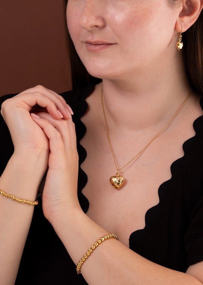 A woman wearing a gold heart pendant necklace, matching gold drop earrings, and a gold chain bracelet, posing against a solid brown background.