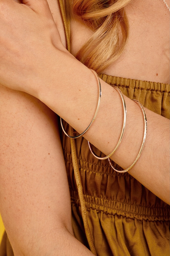 Three thin silver-toned bangles stacked on a woman's wrist, shown in a close-up lifestyle shot with her arm crossed over her chest.