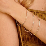 Three thin silver-toned bangles stacked on a woman's wrist, shown in a close-up lifestyle shot with her arm crossed over her chest.