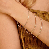 Three thin silver-toned bangles stacked on a woman's wrist, shown in a close-up lifestyle shot with her arm crossed over her chest.