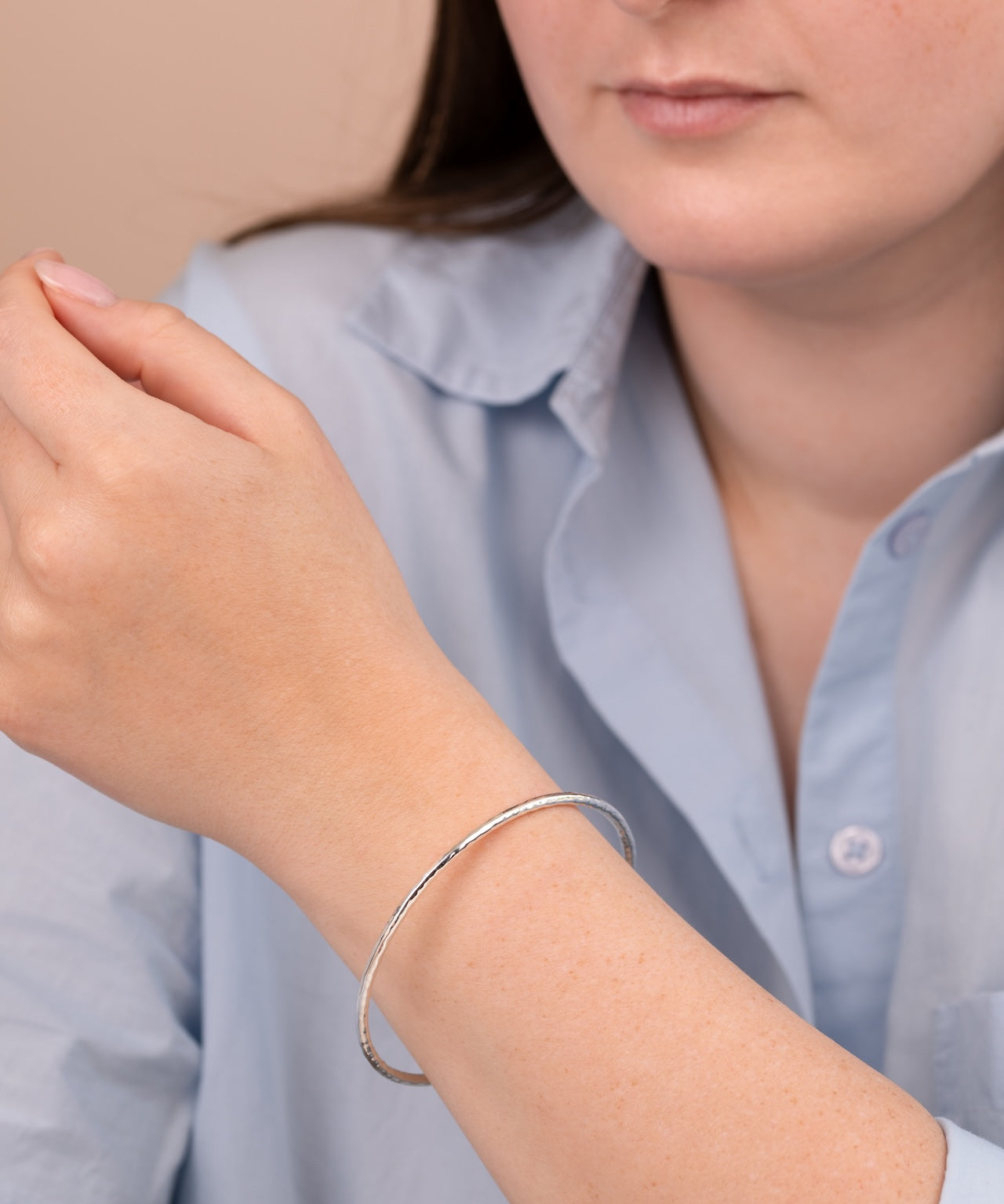 A close-up of a person wearing a thin silver bangle bracelet on their wrist, wearing a light blue button-up shirt against a soft beige background.