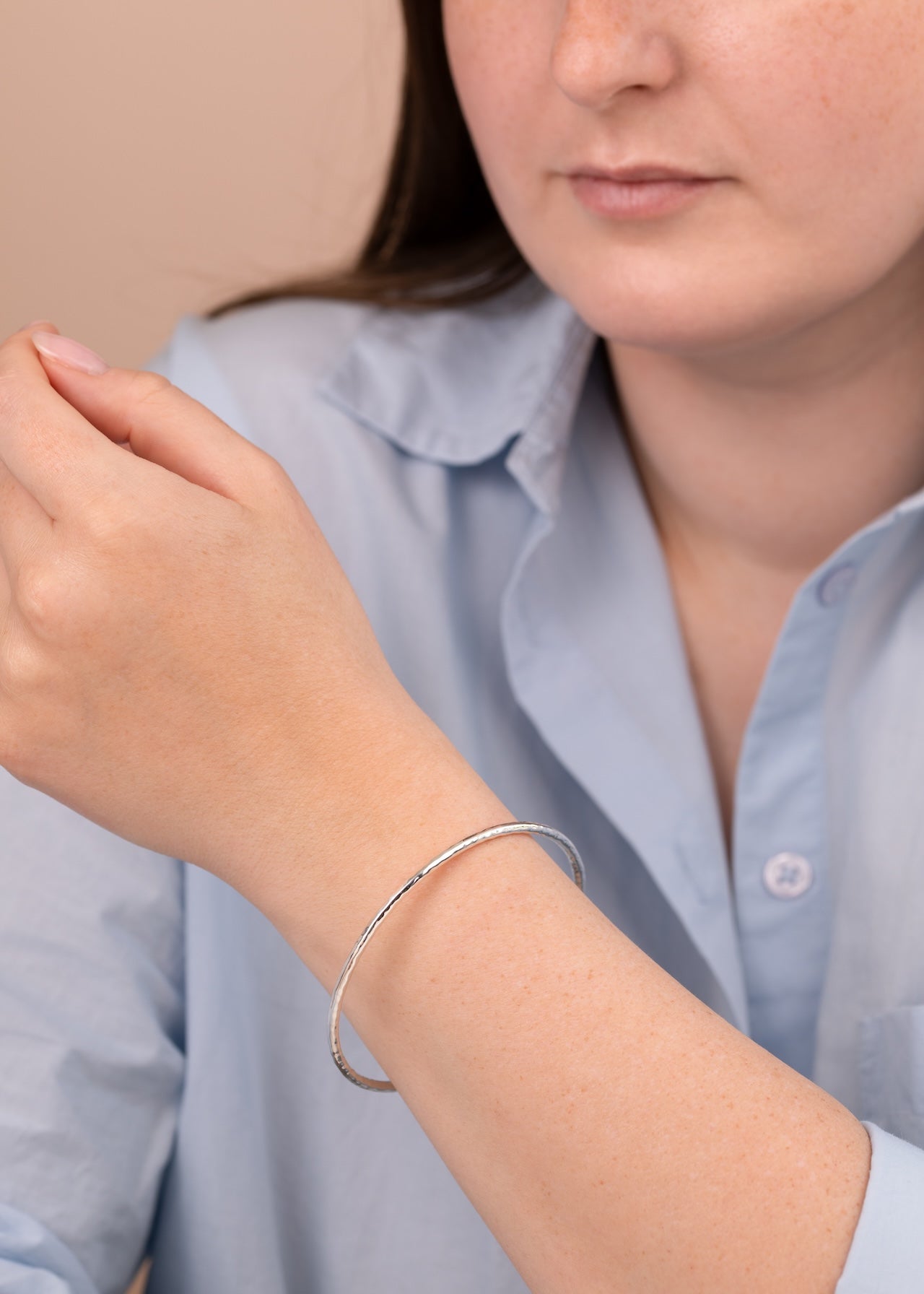 A close-up of a person wearing a thin silver bangle bracelet on their wrist, wearing a light blue button-up shirt against a soft beige background.