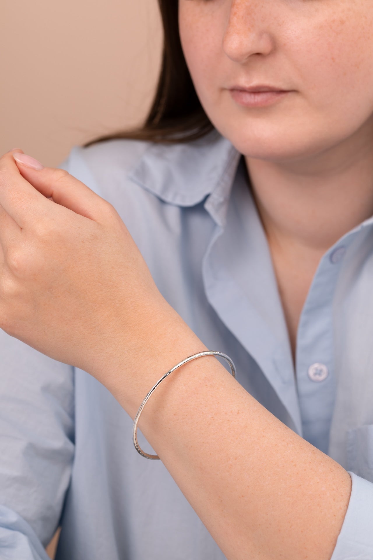 A close-up of a person wearing a thin silver bangle bracelet on their wrist, wearing a light blue button-up shirt against a soft beige background.