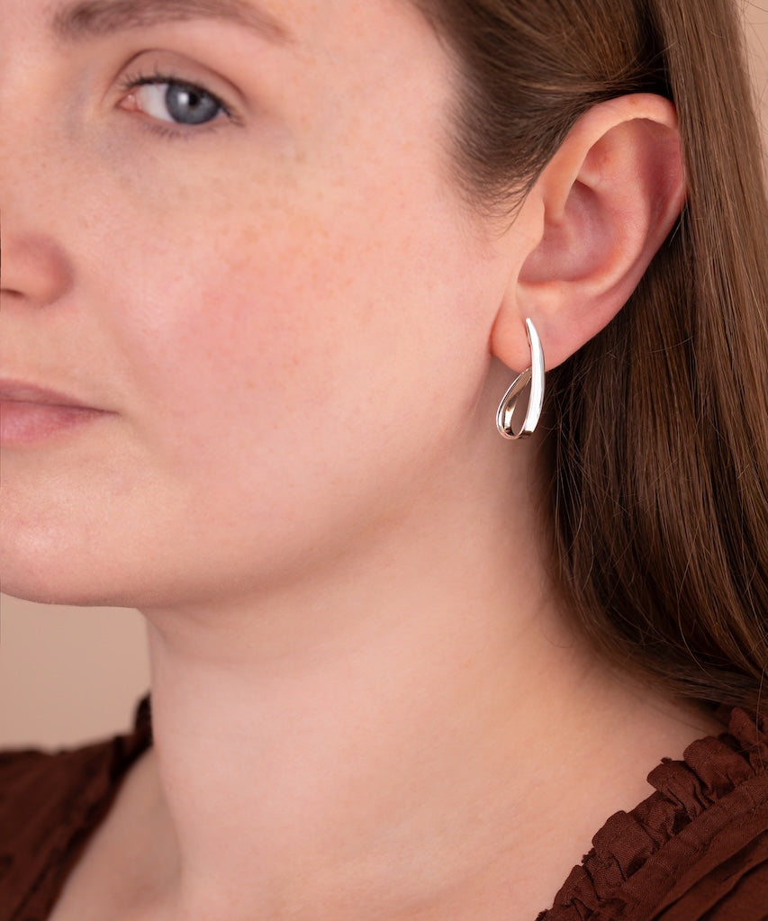 A close-up of a woman wearing a small silver crescent-shaped hoop earring, showing the jewelry on her ear against her face and brown top.