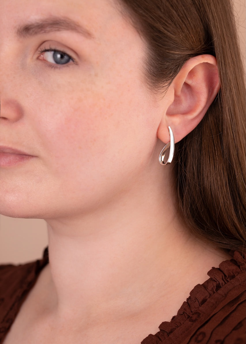 A close-up of a woman wearing a small silver crescent-shaped hoop earring, showing the jewelry on her ear against her face and brown top.