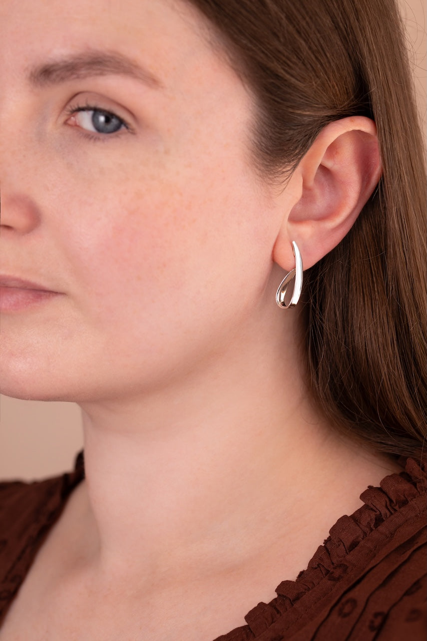 A close-up of a woman wearing a small silver crescent-shaped hoop earring, showing the jewelry on her ear against her face and brown top.