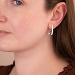 A close-up of a woman wearing a small silver crescent-shaped hoop earring, showing the jewelry on her ear against her face and brown top.