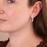 A close-up of a woman wearing a small silver crescent-shaped hoop earring, showing the jewelry on her ear against her face and brown top.