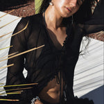 Woman in a black outfit sitting outdoors with sunlight and plants around her.