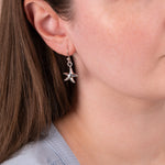 A close-up of a woman wearing a silver dangling earring shaped like a starfish with small clear stones, shown against her ear and neck.