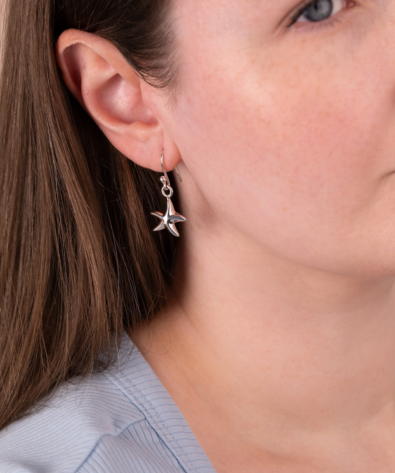 A close-up of a woman wearing a silver dangling earring shaped like a starfish with small clear stones, shown against her ear and neck.