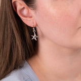 A close-up of a woman wearing a silver dangling earring shaped like a starfish with small clear stones, shown against her ear and neck.