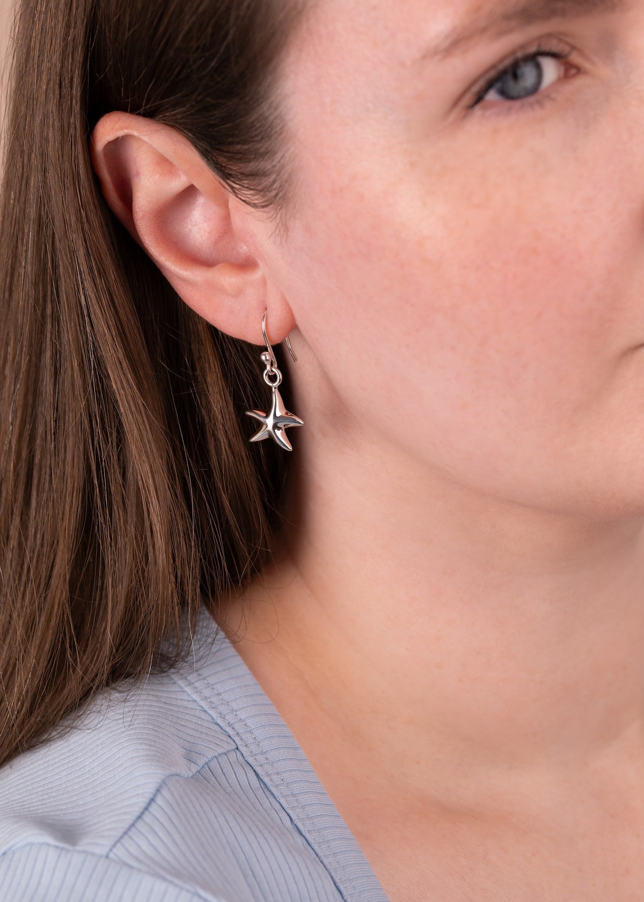 A close-up of a woman wearing a silver dangling earring shaped like a starfish with small clear stones, shown against her ear and neck.