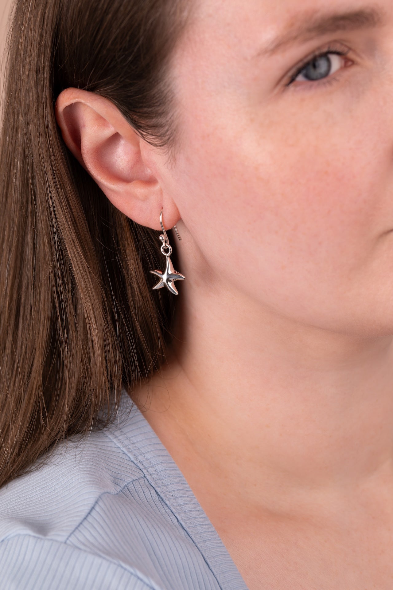 A close-up of a woman wearing a silver dangling earring shaped like a starfish with small clear stones, shown against her ear and neck.
