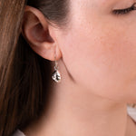 A close-up of a woman's ear wearing a small silver teardrop dangle earring with a clear faceted drop, against her skin and brown hair.