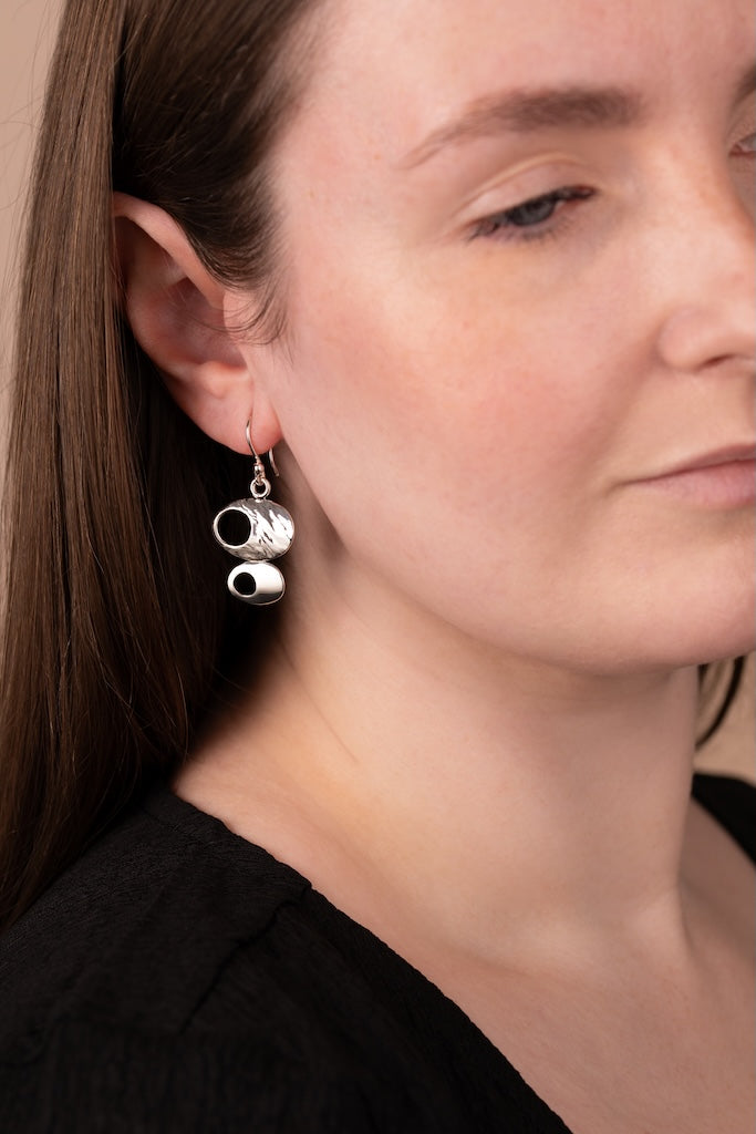 A close-up of a woman wearing a pair of silver dangle earrings shaped like stacked coffee cups, shown against her ear and neck.