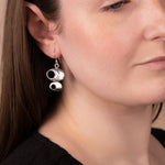 A close-up of a woman wearing a pair of silver dangle earrings shaped like stacked coffee cups, shown against her ear and neck.