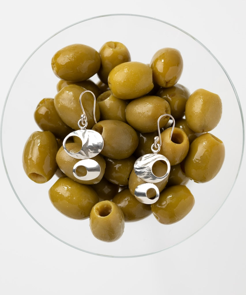 A pair of silver dangle earrings with abstract cutout shapes, displayed on top of green olives inside a clear glass bowl against a light gray background.