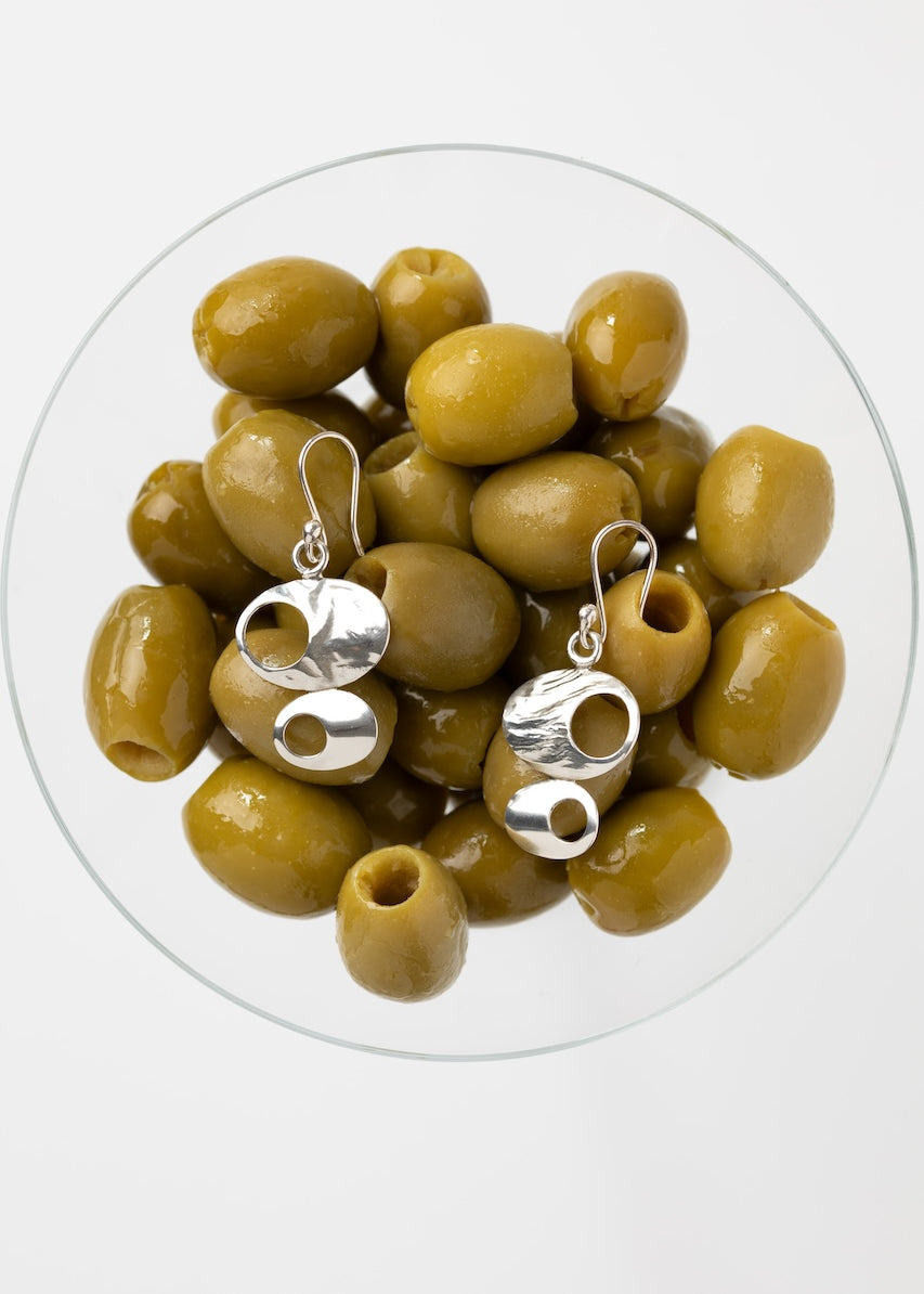 A pair of silver dangle earrings with abstract cutout shapes, displayed on top of green olives inside a clear glass bowl against a light gray background.