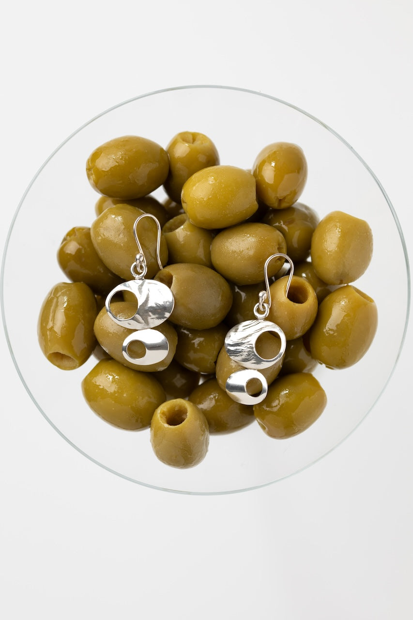 A pair of silver dangle earrings with abstract cutout shapes, displayed on top of green olives inside a clear glass bowl against a light gray background.