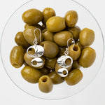 A pair of silver dangle earrings with abstract cutout shapes, displayed on top of green olives inside a clear glass bowl against a light gray background.