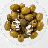 A pair of silver dangle earrings with abstract cutout shapes, displayed on top of green olives inside a clear glass bowl against a light gray background.