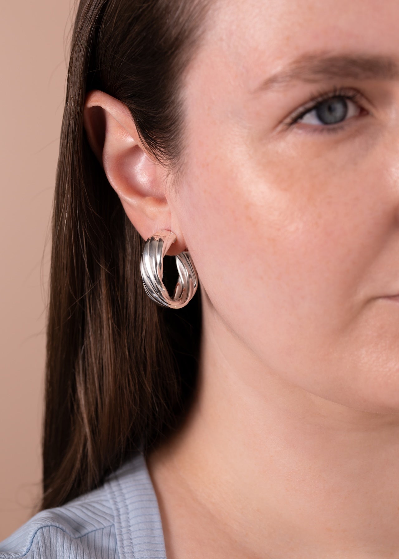 A close-up of a woman wearing a pair of silver twisted hoop earrings, showing the earring on her ear against a solid beige background.