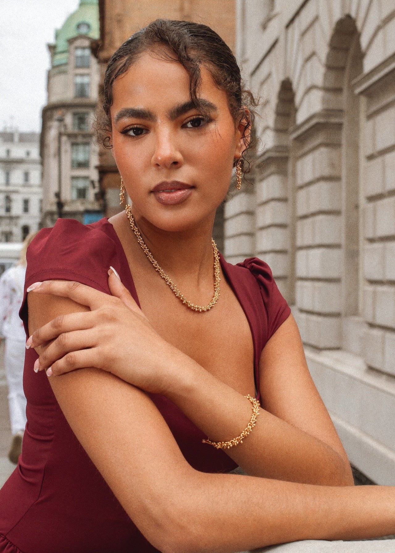 Woman in a burgundy dress with gold jewelry against an urban background